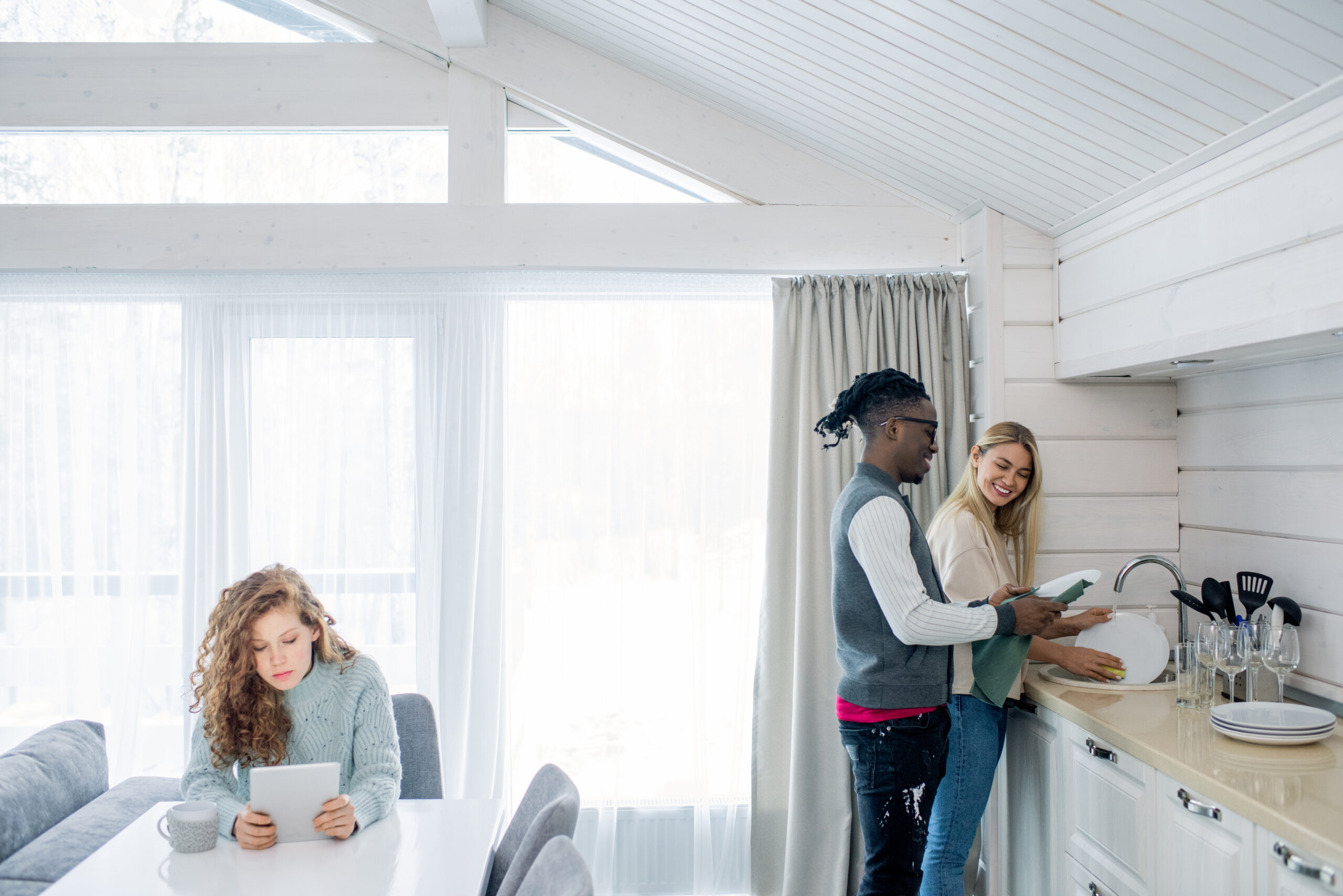 Happy young intercultural couple washing dishes by sink in the kitchen while their friend using mobile gadget by table